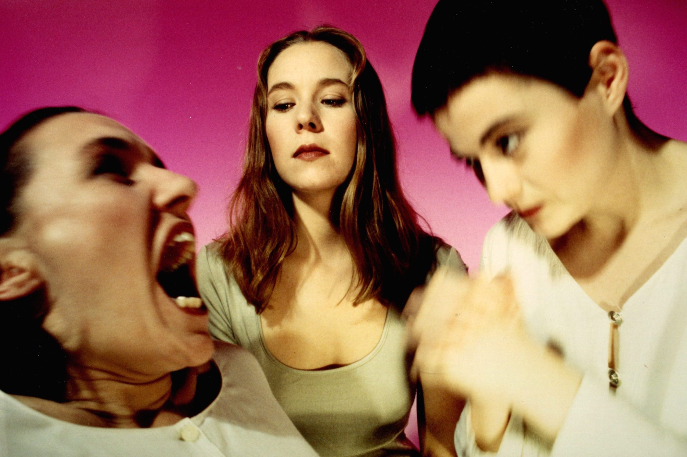 Three woman are stood together against a pink background. 2 women are blurred, one with hteir mouth wide open.