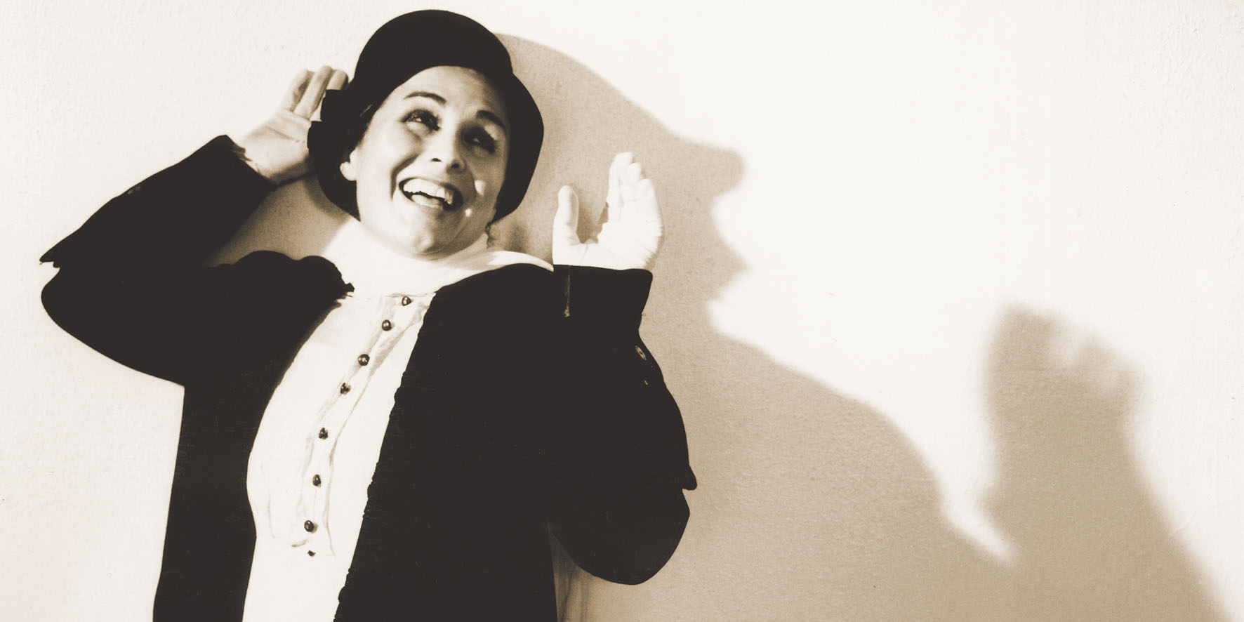 A woman dressed in a white blouse and black top hat smiling with her arms raised against a white background.