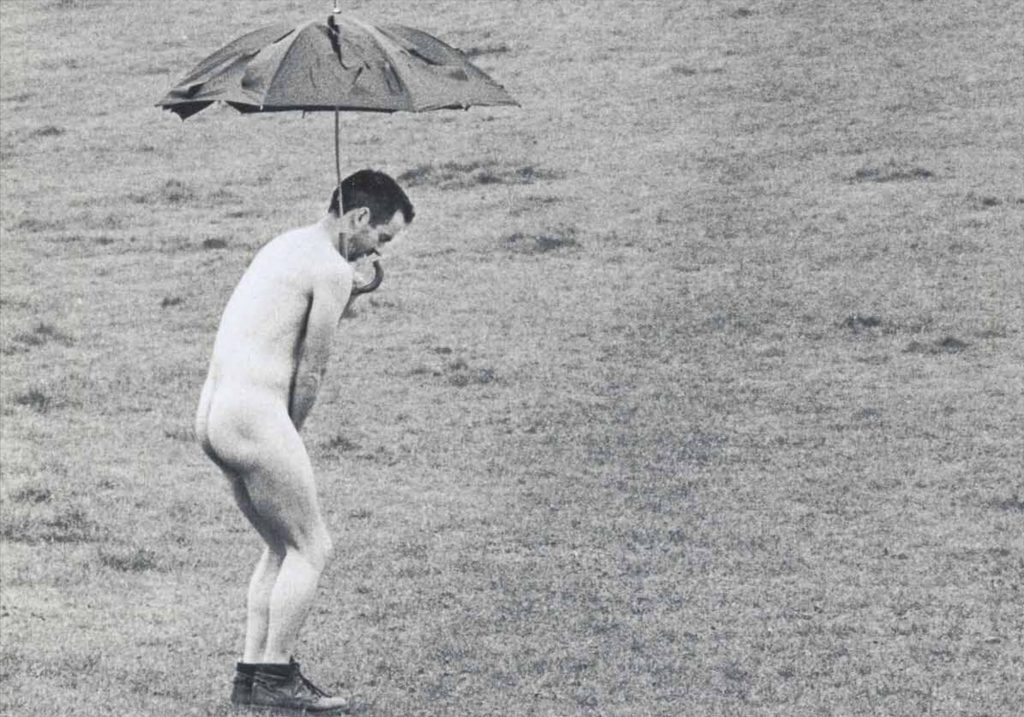 This image displays a picture of a naked man holding an umbrella in open greenery. The image is black and white.