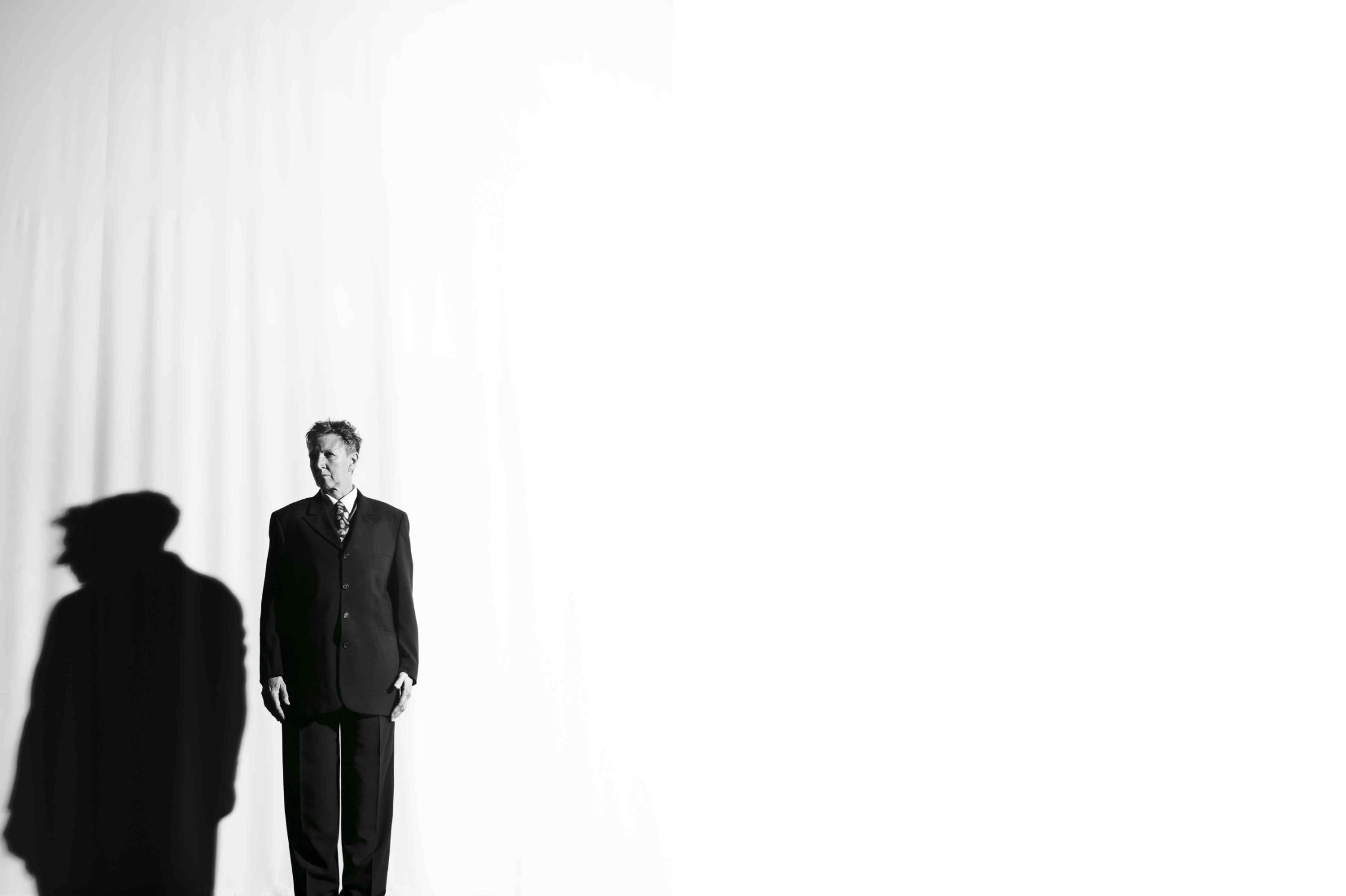 Associate Artist Peggy Shaw dressed in a black suit to the left of the photo looking away from the camera against a white background. Her shadow is seen beside her.