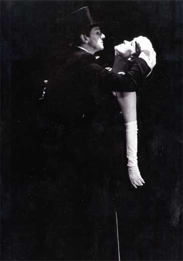 A man holding a woman by the head dressed in a black trench coat and top hat.