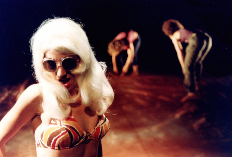 A woman in a blonde wig wearing a bikini. Blurred in the back of the image is two other women facing the floor.