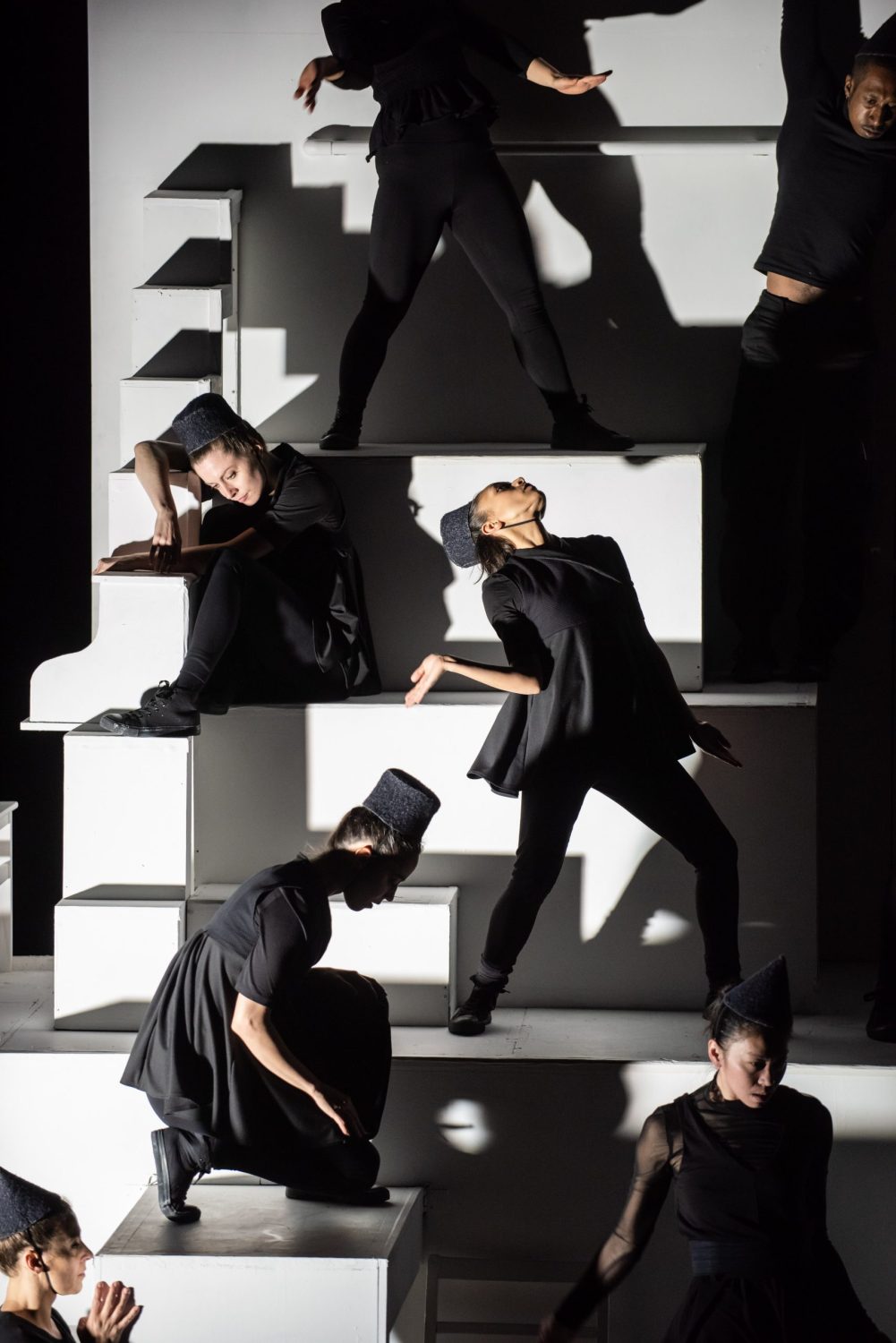 This image displays 7 performers on a white staircase structure identically dressed in black wearing top hats. They are all at different levels and positions.
