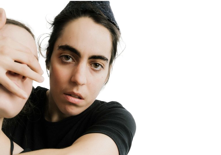 This image displays associate artist Rachele Rapisardi looking into the camera while covering the eyes of another person with her hands. She is against a white background.