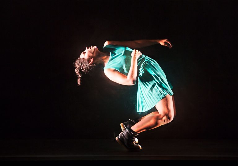 A dancer against a black background with bending backwards towards the ground in a bright green dress