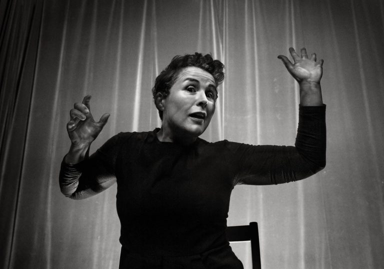 A black and white image with a woman's arms stretched out with her mouth open in front of a draped curtain sitting on a chair.