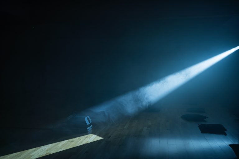 This image displays a beam of light in a dark room, entering from the right and landing on the floor in the bottom left. The room is smoky. The light partially illuminates a smoke machine on the floor. Cushions are dimly visible on the right of the image.
