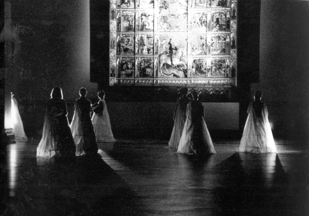 Six women dressed in long open dresses looking up towards a huge picture on the wall. They have their backs to the camera.