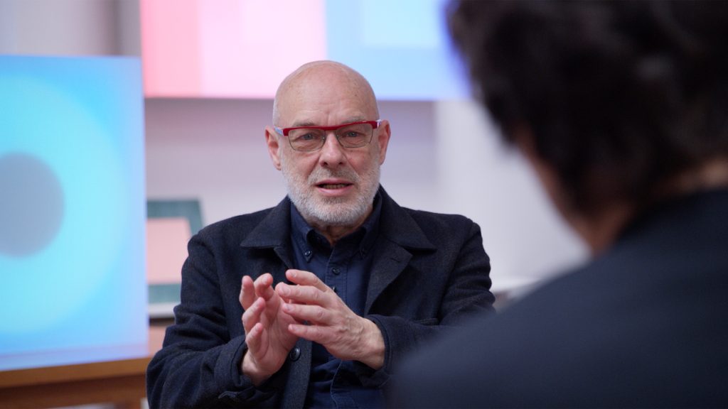 This image displays the musician Brian Eno. He is sitting down and gesturing forwards with both hands. He has a shaved head, a short grey beard, and is wearing glasses.