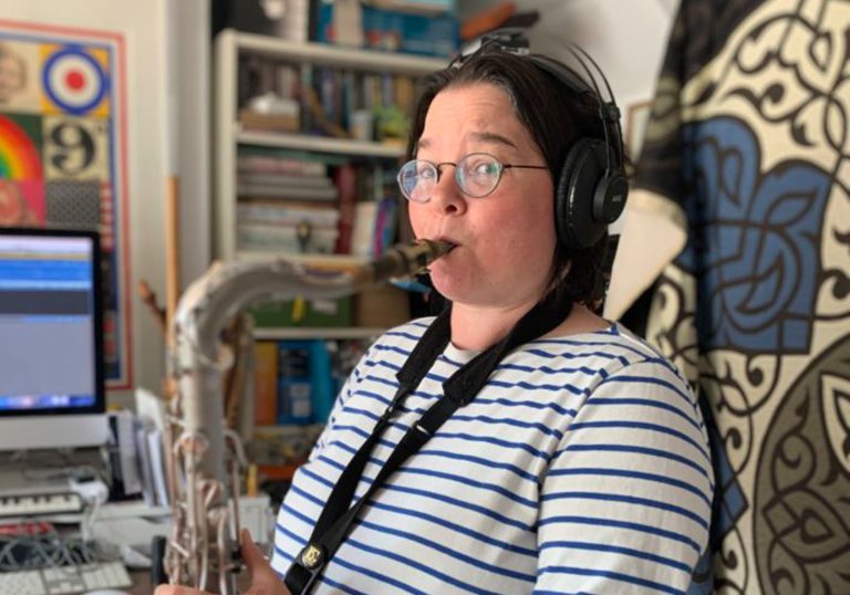A woman in a striped top blowing into a trumpet with a bookshelf behind her