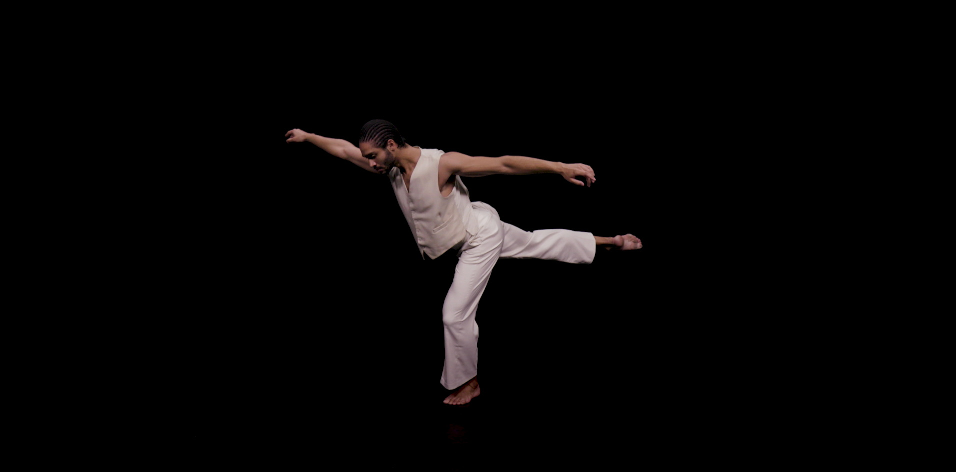 This image displays dancer Harry against a black background with one leg in the air, one arm in front of him and one arm behind him. He is balancing on on leg.