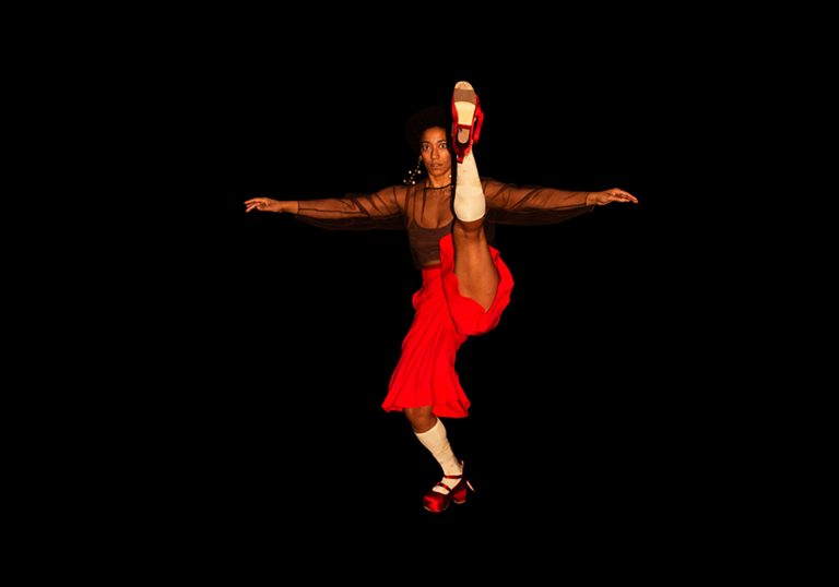 A dancer against a black background with her arms out and one leg in the air.