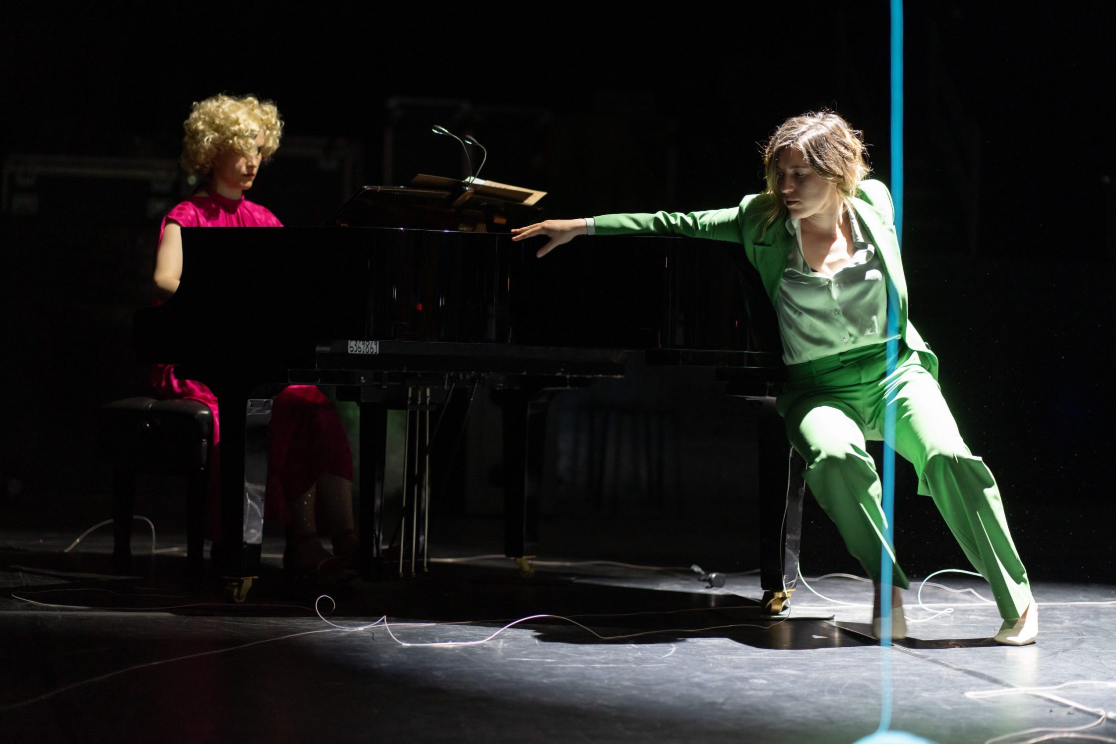 A woman wearing a pastel green suit with her hands stretched holding onto a grand piano. Sitting at the piano is a woman with blonde curly hair wearing a long pink dress.