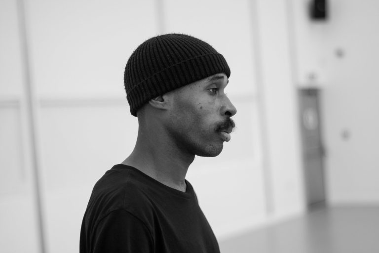 Theo TJ Lowe is pictured from the shoulders up looking off camera so we see his profile. He wearing a black tshirt and black beanie hat, and the blurred background looks like it could be a rehearsal room.