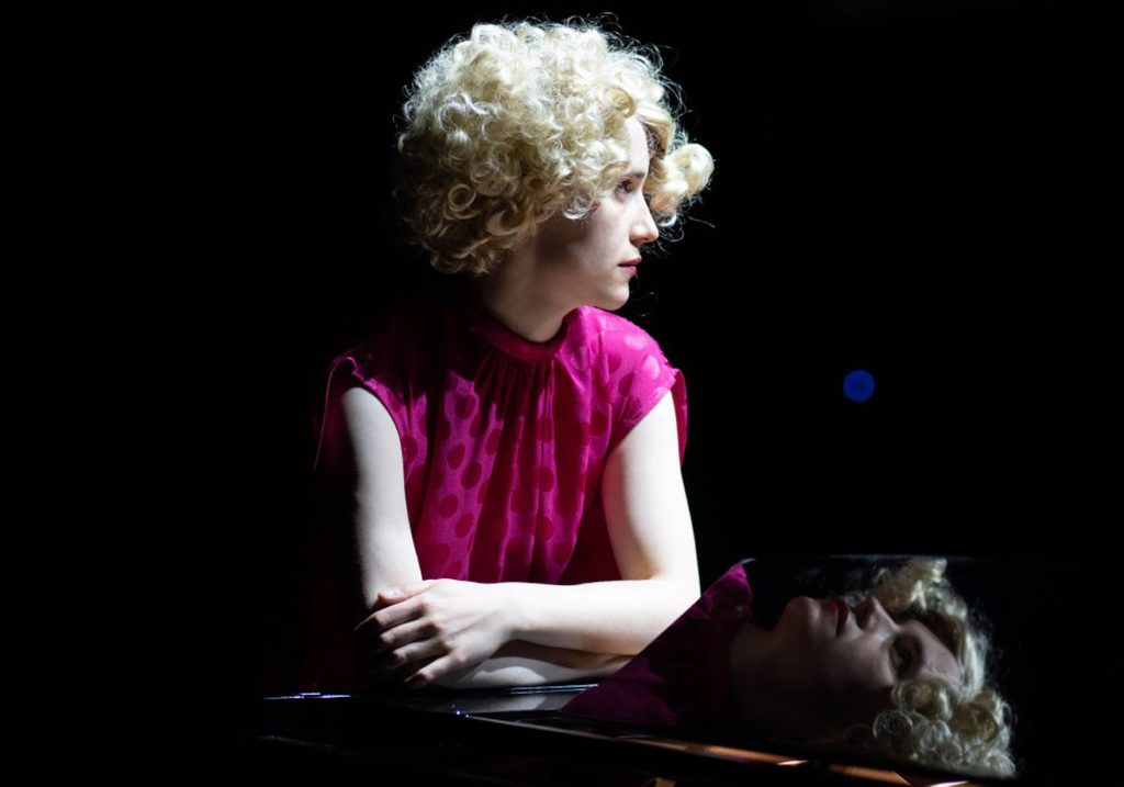 A woman with short blonde curly hair dressed in a pink polka dot blouse looking across from the camera resting her hands on a piano. A reflection of her face is seen in the piano flap.