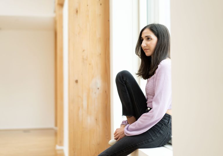 Eliz Hassan posing on a window ledge with one knee bent looking across Clod Ensemble's Skylight Studio. She is wearing a blush pink blouse.