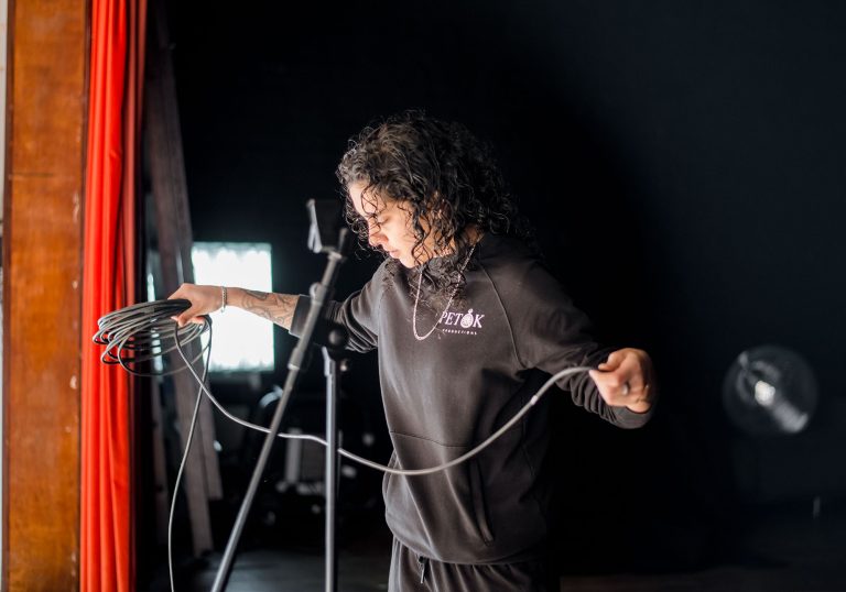 Associate artist Pembe Tokluhan backstage unravelling a wire. A mic stand is placed in front of her. To the left is a bright red stage curtain.