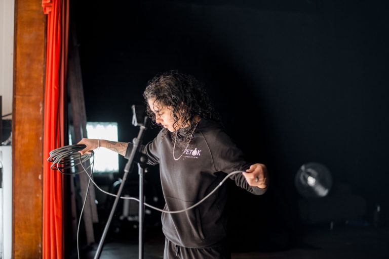A photo of Pembe Tokluhan, the founder of Petok Productions and Clod Ensemble’s Production and Engagement Coordinator. Pembe is looking down to the floor while coiling a microphone cable. The coiled cable is held in her right hand while the end of the cable is held in her left hand. She is facing to the left of the picture, and in front of her we can see a theatre curtain pulled back, meaning she is looking out from the stage to a venue. It is dark behind her. There is a microphone stand before her in the photo, which is out of focus.