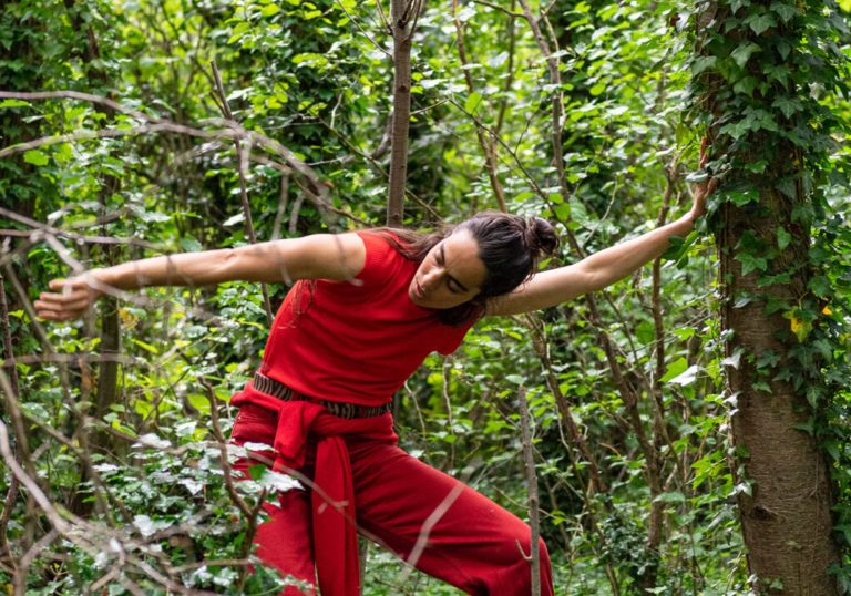 Rachele Rapisardi in a forest with her arms stretched out touching two trees. She is wearing a bright red outfit.
