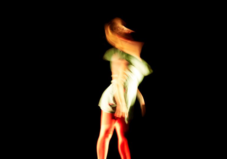 A dancer mid-pose blurred against a plain black background. The dancer is wearing a bright green top and bright red trousers.
