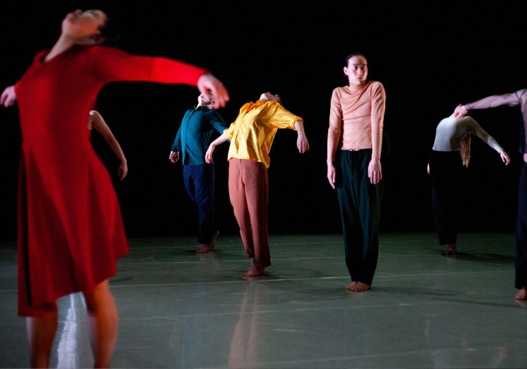 Five dancers standing barefoot on the ground wearing multiple coloured tops. Five dancers have their hands outstretched and one stands in the centre with their hands by their side.