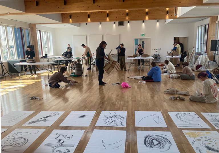 Various drawings aligned across a wooden tile studio floor. Behind the drawings are people people sitting and standing creating sculptures out of cardboard paper.
