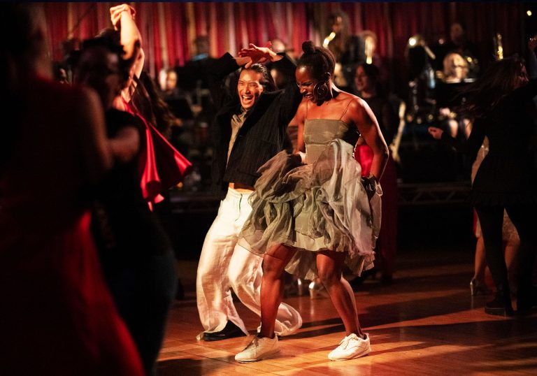 A man and a women laughing whilst dancing in a ballroom setting surrounded by other people dancing and a band behind them.