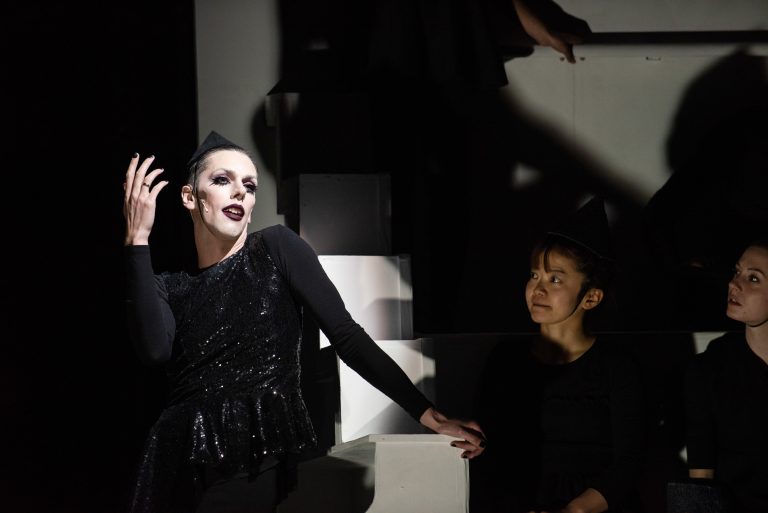 A singer in a black sparkly top with the spotlight on her. One of her hands is leaning on a white block and the other is in the air. She is looking away from the camera. Two people are also in the shot looking at the singer.