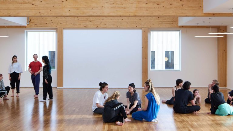 This image depicts a group of people sitting and standing in groups inside a dance studio.