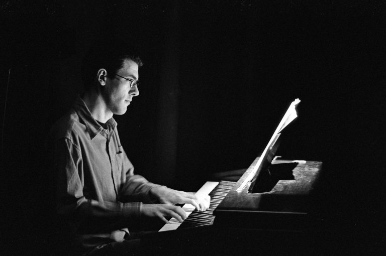 A man playing a piano whilst looking at a musical store placed on a stand on top of the piano. The photo is in black and white.