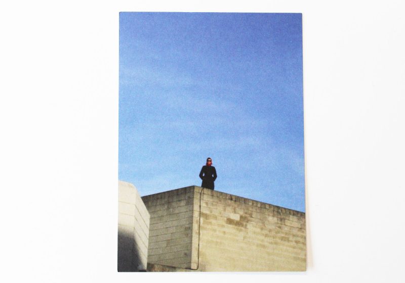 Postcard of a lady against a blue sky standing on top of a building.