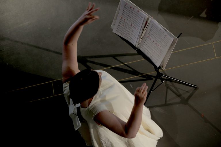 A woman has her hands raised looking at a musical score which is placed in front of her on a music stand. She is wearing a cream dress.