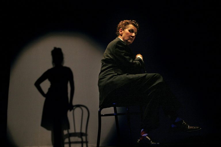Associate Artist Peggy Shaw looking towards the camera in a black suit. There is a silhouette of a woman. holding a chair behind her.
