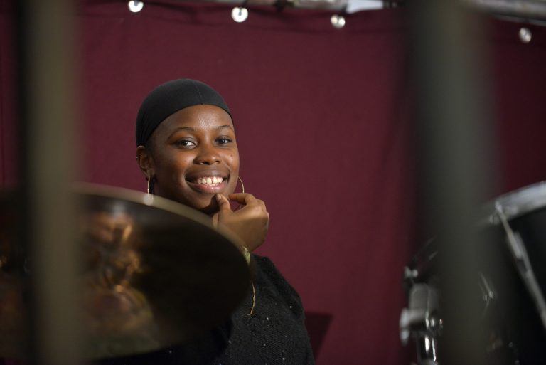 Associate artist Vanessa smiling looking into the camera. Blurred to the right of her is a drumkit.