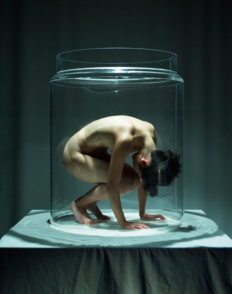A naked woman under a glass vitrine on a table covered by a tablecloth. Her head is low, knees bent and arms reached out.