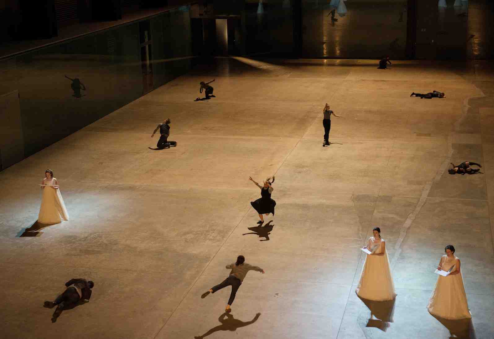 Image of several people in a turbine hall spread out in different positions and heights. 3 people are dressed in white ball gowns holding an opened book.
